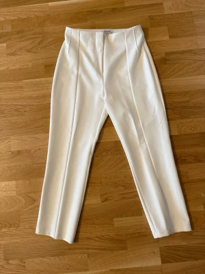 Vita kostymbyxor från Klapa - Snygga vita kostymbyxor från Klapa med hög midja och pressveck framtill. Byxorna har raka ben och diskret dragkedja i sidan. Perfekta för en clean och stilren look. Materialet känns mjukt och har en lätt stretch för extra komfort.