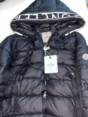Moncler cardere Jacka - 1500 men kan gå ner i pris. Snygg svart dunjacka från Moncler med stor huva och dragkedja framtill. Jackan har två framfickor med dragkedja, justerbar dragsko i huvan och Moncler-logga på ärmen. Tillverkad i glansigt material som ger en modern look.