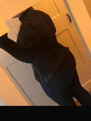 Svart topp med mönster och volang - Unik svart kofta med stora beige mönster framtill och dekorativ volangkant nedtill. Koftan har knäppning fram och är långärmad. 