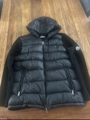Svart hybrid cardiganjacka Moncler - Snygg svart cardiganjacka från Moncler med vadderad framsida och huva i blankt material. Ärmar och rygg är stickade i mjuk ullmix. Två dragkedjefickor fram och Moncler-logga på ärmen. Kom görna med prisförslag eller frågor.😊