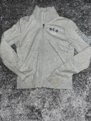 Grå zip hoodie från Hollister - Snygg grå zip hoodie från Hollister med broderad text på bröstet och ärmen. Tröjan har hög krage, två fickor fram och dragkedja hela vägen. Tillverkad i mjukt bomullsmaterial som är perfekt för chill dagar. Klassisk och enkel design med Hollister-logga. Storlek L (barn)