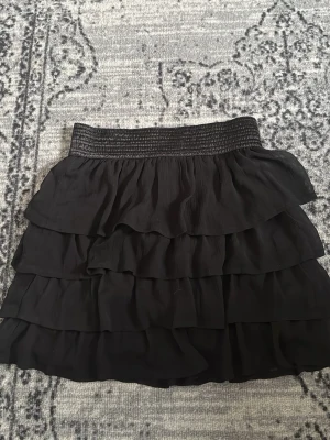 Svart volangkjol med dragkedja - Kort svart kjol med flera lager volanger och bred resår i midjan. Kjolen har dragkedja bak och är gjord i ett lätt, flowy material som ger en snygg siluett. Perfekt för dig som gillar en trendig och feminin stil.