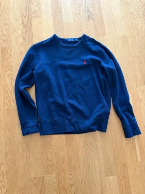 Mörkblå sweatshirt från Polo Ralph Lauren - Klassisk mörkblå sweatshirt från Polo Ralph Lauren med rund halsringning och röd broderad logga på bröstet. Tröjan har ribbade muddar vid ärmslut och nederkant samt en snygg detaljsöm vid halsen. Perfekt för en avslappnad och stilren look.