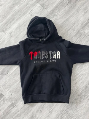 Svart Trapstar hoodie med brodyr - Säljer en svart hoodie från Trapstar med stor broderad logga i rött, grått och vitt på bröstet samt texten 'IT'S A SECRET' under. Hoodien har en klassisk huva med dragsko och en stor magficka. Storlek S