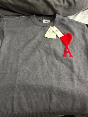 Grå stickad tröja från Ami Paris - Snygg grå stickad tröja från Ami Paris med rund halsringning och ribbade muddar. På bröstet finns ett stort rött hjärta med bokstaven A. Perfekt för dig som gillar stilrena detaljer och vill ha något extra i garderoben.