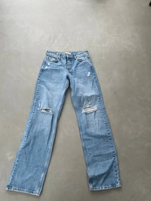 Ljusblå ripped jeans från Zara - Säljer ett par ljusblå raka jeans från Zara med slitningar och hål på båda benen. Jeansen har klassisk femficksmodell och är tillverkade i denim. Perfekta för en avslappnad och trendig look.