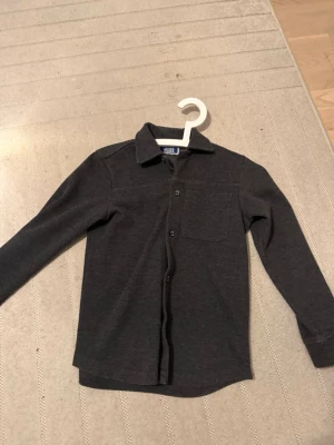 Mörkgrå skjorta från Jack & Jones ny pris 800kr - Stilren mörkgrå skjorta från Jack & Jones med klassisk krage, bröstficka och knäppning framtill. Skjortan har lång ärm och är tillverkad i mjukt material som känns bekvämt mot huden. Perfekt för en avslappnad men snygg look.