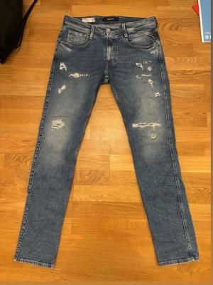 Blå slitna jeans från Replay - Säljer ett par blå jeans från Replay med slim fit och slitningar framtill för en trendig look. Jeansen har klassisk femficksmodell, dragkedjegylf och snygga detaljer på bakfickorna. Perfekta för dig som gillar en avslappnad och modern stil. Hör av er med frågor och funderingar!🙌