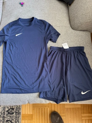 Mörkblått Nike träningsset - Säljer ett mörkblått träningsset från Nike med t-shirt och shorts. Båda plaggen har den klassiska vita Nike-loggan och är gjorda i ett lätt och snabbtorkande material, perfekt för träning eller sportaktiviteter.