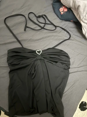 Svart bandeau topp med hjärtdetalj - Snygg svart bandeau topp med tunna axelband och en söt hjärtformad detalj. Perfekt för dig som vill ha en trendig och stilren look. Passar till många olika outfits och är enkel att matcha.