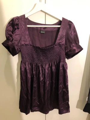 Lila puffärmad topp  - Supersöt lila topp med puffärmar och smock över bysten. Tillverkad i glansigt material som ger en lyxig känsla. Fyrkantig ringning och lös passform nedtill. 