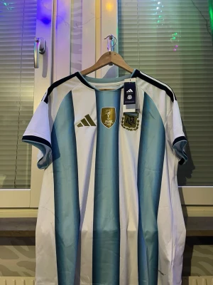 Argentina matchtröja Adidas blåvit 2022 storlek L - Säljer en officiell Argentina matchtröja från Adidas med klassiska blå och vita ränder. Tröjan har guldfärgade detaljer, FIFA World Champions 2022-märke och AFA-logga på bröstet. Tillverkad i lätt och ventilerande material, perfekt för fotboll eller träning.