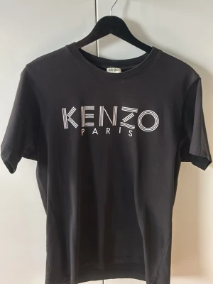 Svart Kenzo t-shirt med logga - Snygg svart t-shirt från Kenzo med stor vit logga 'KENZO PARIS' på bröstet. Klassisk rund halsringning och korta ärmar. Tillverkad i mjuk bomull som känns skön mot huden. Perfekt för dig som gillar stilrena och trendiga plagg.