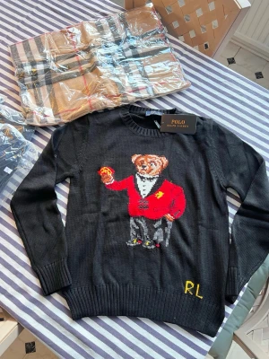 Svart stickad tröja Polo Ralph Lauren - Svart stickad tröja från Polo Ralph Lauren med ikonisk björn i röd kavaj broderad på framsidan. Rund halsringning och ribbade muddar. Detaljer i gult och grått samt RL-broderi nertill. Perfekt för dig som gillar statement-plagg.