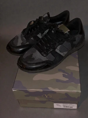 Valentino Rockrunner camo sneakers - Snygga Valentino Rockrunner sneakers i svart och grått camouflagemönster. Skorna har snörning, rund tå och platt sula. Materialmix med textil och skinn, samt detaljer i svart. Perfekta för dig som gillar streetstyle och exklusiva märken. Skriv vid frågor 