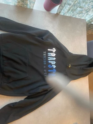 Svart Trapstar hoodie med tryck - Snygg svart hoodie från Trapstar med stor magficka och huva. Framsidan har ett fett vitt och blått Trapstar-tryck samt texten 'It's A Secret' i mindre bokstäver. Hoodien har guldiga öljetter vid snörningen och är gjord i mjukt bomullsmaterial.