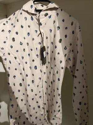 Mönstrad skjorta från Oscar Jacobson - Säljer en ljus skjorta från Oscar Jacobson med blå och gröna prickar. Skjortan har lång ärm, klassisk krage och knäppning framtill. Slim fit-modell i mjuk bomull, perfekt för dig som gillar stilrena men lekfulla detaljer.