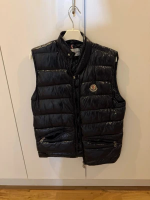 Svart dunväst från Moncler - Snygg svart dunväst från Moncler med glansig finish och ståkrage. Västen har två dragkedjeförsedda fickor framtill och klassisk Moncler-logga på bröstet. Perfekt för lager-på-lager och streetstyle. Materialet är dunfyllning med ett yttertyg i nylon.