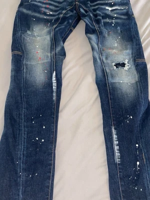 MörkblåDsquared2 jeans med färgstänk - Säljer ett par mörkblå jeans med slitningar, patchdetaljer och vita samt röda färgstänk för en cool och unik look. Jeansen har kontrastsömmar och klassisk femficksmodell. Perfekta för dig som gillar streetwear och vill sticka ut. (storlek för 165-175)