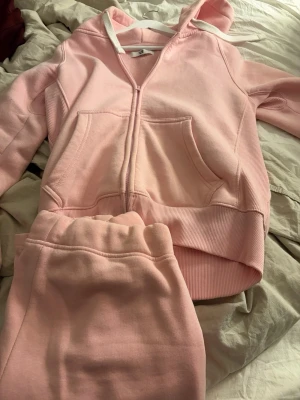 Rosa loungewear-set från russemerch  - Mysigt loungewear-set i ljusrosa från russemerch Setet består av en hoodie med dragkedja, fickor och vit dragsko samt matchande mjukisbyxor med resår och dragsko i midjan. Perfekt för chill dagar hemma. I storlek s i byxorna och m i tröjan den har en liten fläck på sig men inget annat 
