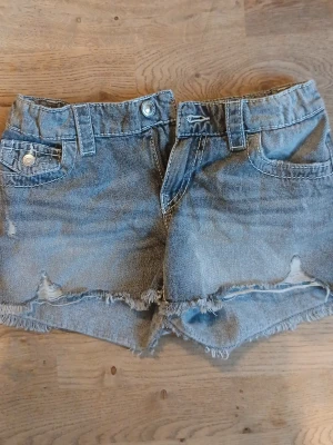 Gråa jeansshorts med broderad ficka - Säljer ett par blå jeansshorts med fransig kant och snygg broderad bakficka. Shortsen har normal passform, bälteshällor och klassiska fem fickor. Perfekta för sommaren och ger en avslappnad look.