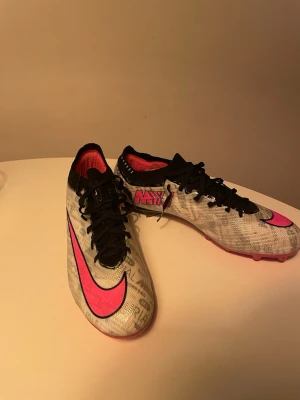 Nike Mercurial fotbollsskor vit/rosa - Snygga Nike Mercurial fotbollsskor i vitt med svarta snören och detaljer samt stora rosa swoosh-loggor. Perfekta för dig som vill sticka ut på matchen. Säljer de då jag tyckte de var för små. Använda 4 träningar enbart. 
