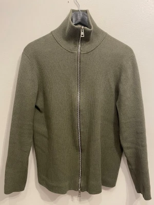 Olivgrön ribbad ziptröja från Ljung - Snygg olivgrön ribbstickad tröja från Ljung by Marcus Larsson. Tröjan har hög krage och dragkedja framtill, vilket ger en clean och modern look. Perfekt till lager-på-lager och enkel att matcha med olika outfits.