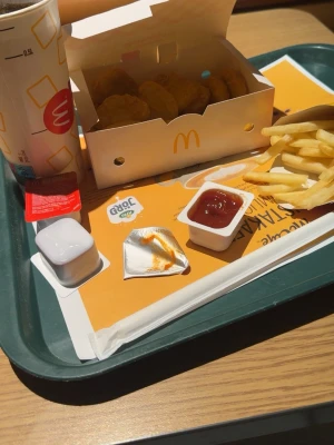 McDonald's menybricka med nuggets - En klassisk McDonald's menybricka med kycklingnuggets i vit kartong, pommes frites, läsk i pappersmugg och flera såser. Allt serveras på en grön bricka med papper under. Perfekt för dig som gillar snabbmat och vill ha en komplett måltid.