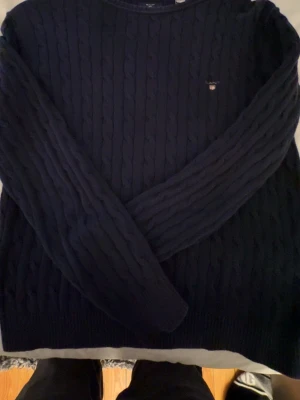 Mörkblå kabelstickad tröja från GANT - Snygg mörkblå kabelstickad tröja från GANT med rund halsringning och diskret logga broderad på bröstet. Tröjan har långa ärmar och ribbade muddar vid ärmslut och nederkant. Perfekt nu till våren- nypris 800