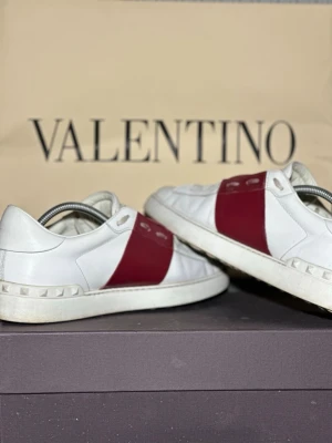 Valentino open - Snygga vita sneakers från Valentino med en bred röd läderdetalj över mitten och ikoniska nitar längs sulan. Skorna har en klassisk låg profil och är tillverkade i läder för en lyxig känsla. Perfekta för dig som vill sticka ut med stilrena detaljer. Og ingår ej