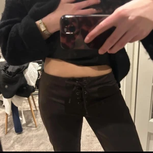 Bruna mocka byxor med snörning fram - Säljer ett par bruna byxor med låg midja och snygg snörning framtill. De har en åtsittande passform och dragkedja i sidan för enkel påklädning. Perfekta för dig som vill ha en trendig och bekväm look.