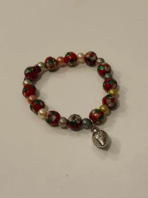 Armband med jordgubbshänge - Armband med röda, gröna och guldiga pärlor i metall och plast, dekorerade med blommönster och små färgskiftningar. Armbandet har ett silverfärgat jordgubbshänge som detalj.