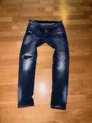 Mörkblå distressed jeans från Dsquared2 - ‼️ÄKTA!!  MÅSTE SY LAPPEN SOM GÖR HÅLET ATT INTE VISA BENET‼️Säljer ett par mörkblå jeans från Dsquared2 med slitningar, färgstänk och en slim fit passform. Jeansen har klassisk femficksdesign, låg midja och detaljerade slitningar på benen för en trendig look. Perfekta för dig som gillar streetwear och edgy stil.