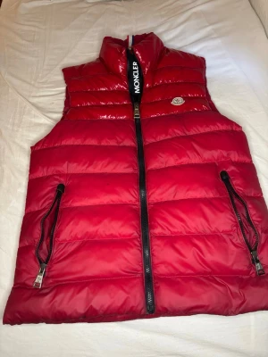 Röd dunväst från Moncler - Snygg röd dunväst från Moncler med hög krage och svart dragkedja framtill. Västen har två fickor med dragkedja och Moncler-logga på bröstet. Tillverkad i glansigt material och har en quiltad design som ger en modern look.