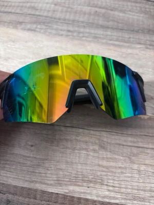 Sportiga solglasögon med spegelglas - Säljer ett par sportiga solglasögon med stora, spegelblå och gröna linser. Bågarna är svarta och har en modern, aerodynamisk design som passar perfekt för utomhusaktiviteter. Snyggt och futuristiskt utseende som sticker ut.