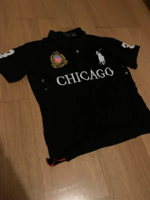 Svart pikétröja Polo Ralph Lauren - Svart pikétröja från Polo Ralph Lauren med stor vit logga på bröstet, broderad Chicago-text och emblem. Klassisk krage och knappar framtill. Snygga detaljer på ärmarna och broderade märken ger en sportig vibe. Storlek M men sitter som S 
