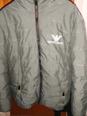 Grön pufferjacka från Emporio Armani - Snygg grön pufferjacka från Emporio Armani med diskret mönster i tyget och logga tryckt på bröstet. Jackan har dragkedja framtill, två fickor med knappstängning och hög krage. Perfekt för kalla dagar och ger en clean streetwear-look.
