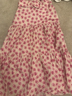 Söt Långkjol - Säljer en ljusrosa långkjol med rosa blommönster och volanger. Kjolen har ett matchande tygbälte med rund spänne i midjan och är perfekt för somriga dagar. Den har en luftig och flowy passform som ger en romantisk vibe. Bältet går att ta av