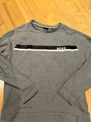 Grå sweatshirt från BOSS - Snygg grå sweatshirt från BOSS med svart och vit rand över bröstet och BOSS-logga. Tröjan har rund halsringning och långa ärmar. Perfekt för en avslappnad och stilren look.