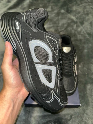 Dior  - Säljer ett par svarta Dior sneakers med reflekterande silverdetaljer och tydlig Dior-logga på sidan. Skorna har en chunky sula, meshpaneler för extra andningsförmåga och svarta skosnören. Perfekta för dig som vill sticka ut med en modern och sportig look.