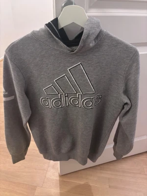 Grå hoodie från Adidas med logga - Snygg grå hoodie från Adidas med stor vit logga på bröstet och huva. Tillverkad i mjukt sweatshirtmaterial som är perfekt för chill dagar. Klassisk passform med ribbade muddar vid ärmar och nederkant. Adidas-detalj även på ärmen.fattas dock snöret därav priset men klassiker 