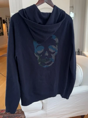 Svart hoodie med dödskalletryck - Mörkblå hoodie från Zadig & Voltaire med dragkedja, huva och snörning. Stor dödskalle i blått och grönt på ryggen och diskret ZV-logga på bröstet. Texten 'MAJOR RESOURCE LEGACY' längs ena ärmen. Mjuk och skön, perfekt för en cool streetstil.