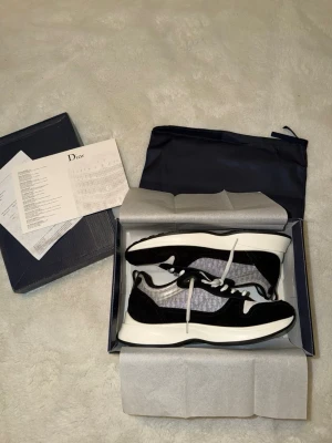 Dior sneakers i svart och vitt - Snygga Dior sneakers i svart och vitt med klassisk monogramdetalj på sidan. Skorna har vita snören, svart mocka och meshpaneler för en modern look. Tjock vit sula ger extra komfort och stil. Perfekta för dig som vill sticka ut med exklusiva detaljer.