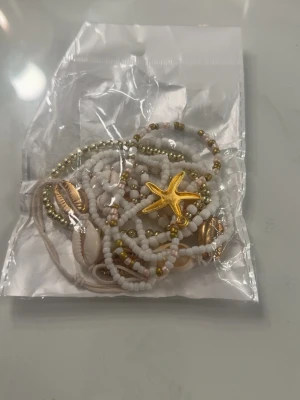 Armband med pärlor och snäckor - Flera armband med vita och guldiga pärlor, dekorerade med snäckor och en guldfärgad sjöstjärna. Armbanden har strandig vibe och mixar olika material som plastpärlor och snäckskal. Perfekt för dig som gillar boho-stil och detaljer.