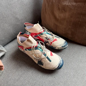 Puma fotbollsskor (future 7 ultimate wave FG/AG) - Unika fotbollsskor från Puma med beige bas och färgstarkt japanskt inspirerat mönster i rött, blått och vitt. Skorna har snörning, låg profil och mönstrad ovandel i syntetmaterial. Perfekta för dig som vill sticka ut på planen. Säljer pga för små, aldrig använda. Nypris 2899kr. Passform 43-45