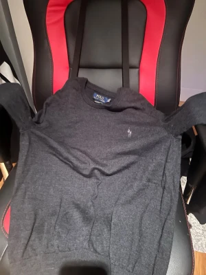 Mörkgrå tröja från Polo Ralph Lauren - Mörkgrå rundhalsad tröja från Polo Ralph Lauren med den klassiska broderade loggan på bröstet. Tröjan har en enkel och stilren design med ribbade muddar vid ärmslut och nederkant. Perfekt för dig som gillar en clean och tidlös look.