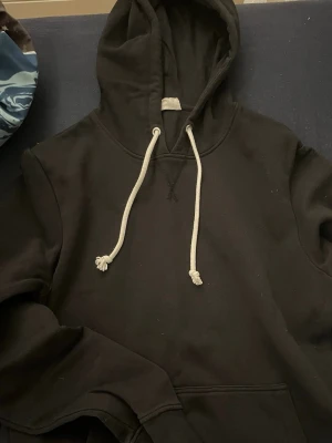 Svart hoodie med vita snören - En klassisk svart hoodie med stora fickor framtill och vita dragsnören i huvan.