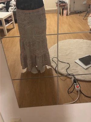 långkjol med paisleymönster - Säljer en ljusblå, grön långkjol med paisleyinspirerat mönster i beige och rosa toner. Kjolen har resår i midjan och volang längst ner för en bohemisk vibe. Perfekt för dig som gillar flowiga och bekväma plagg.