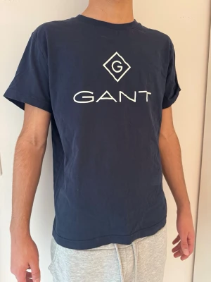 Mörkblå GANT t-shirt  - Mörkblå t-shirt från GANT med stor vit logga och tryck på bröstet. Klassisk rak passform och rund halsringning. Tillverkad i mjuk bomull som känns skön mot huden. Har storlek XS-S. Står L men är L i barnstorlek 