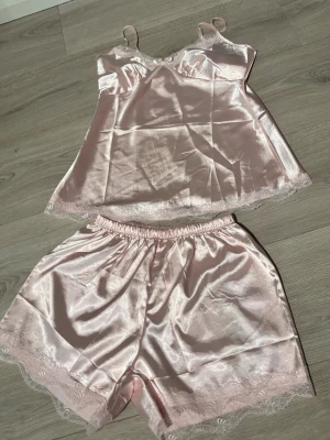 Ljusrosa satinpyjamas med spetsdetaljer - Söt ljusrosa pyjamas i glansigt satinmaterial med linne och shorts. Linnet har tunna axelband, spetskant och en liten rosett framtill. Shortsen har elastisk midja och spetsdetaljer vid bensluten. Perfekt för mysiga kvällar hemma.